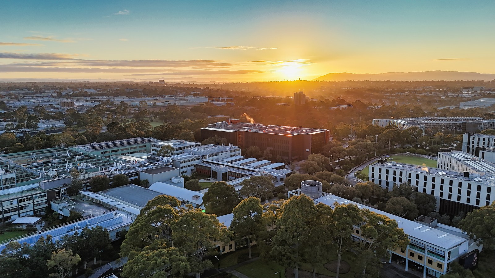 A broad elevated view of buildings and trees beneath a golden sunset and distant hills.