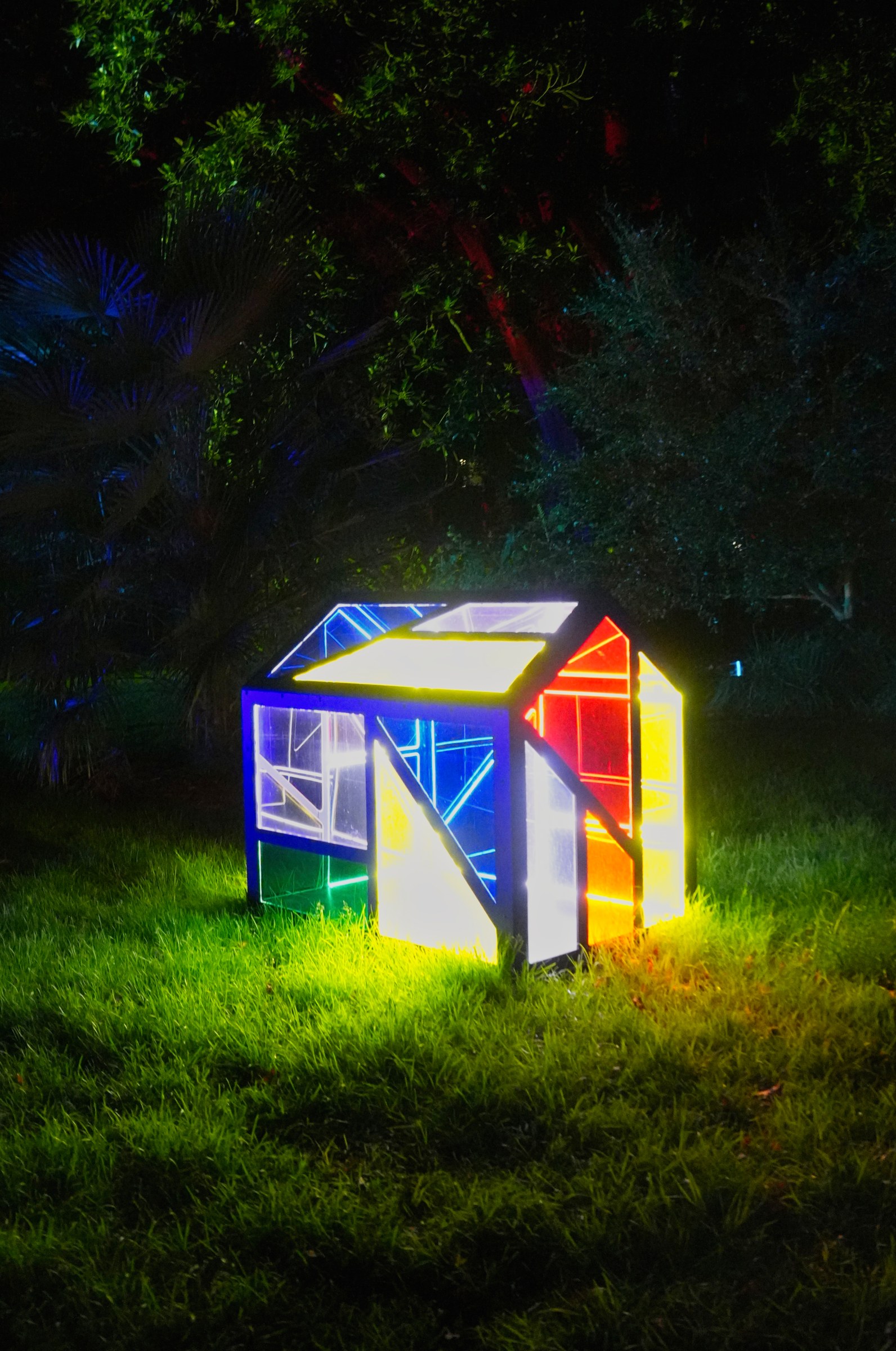 A small geometric structure glows in blue, yellow and red on grass beneath dark trees.