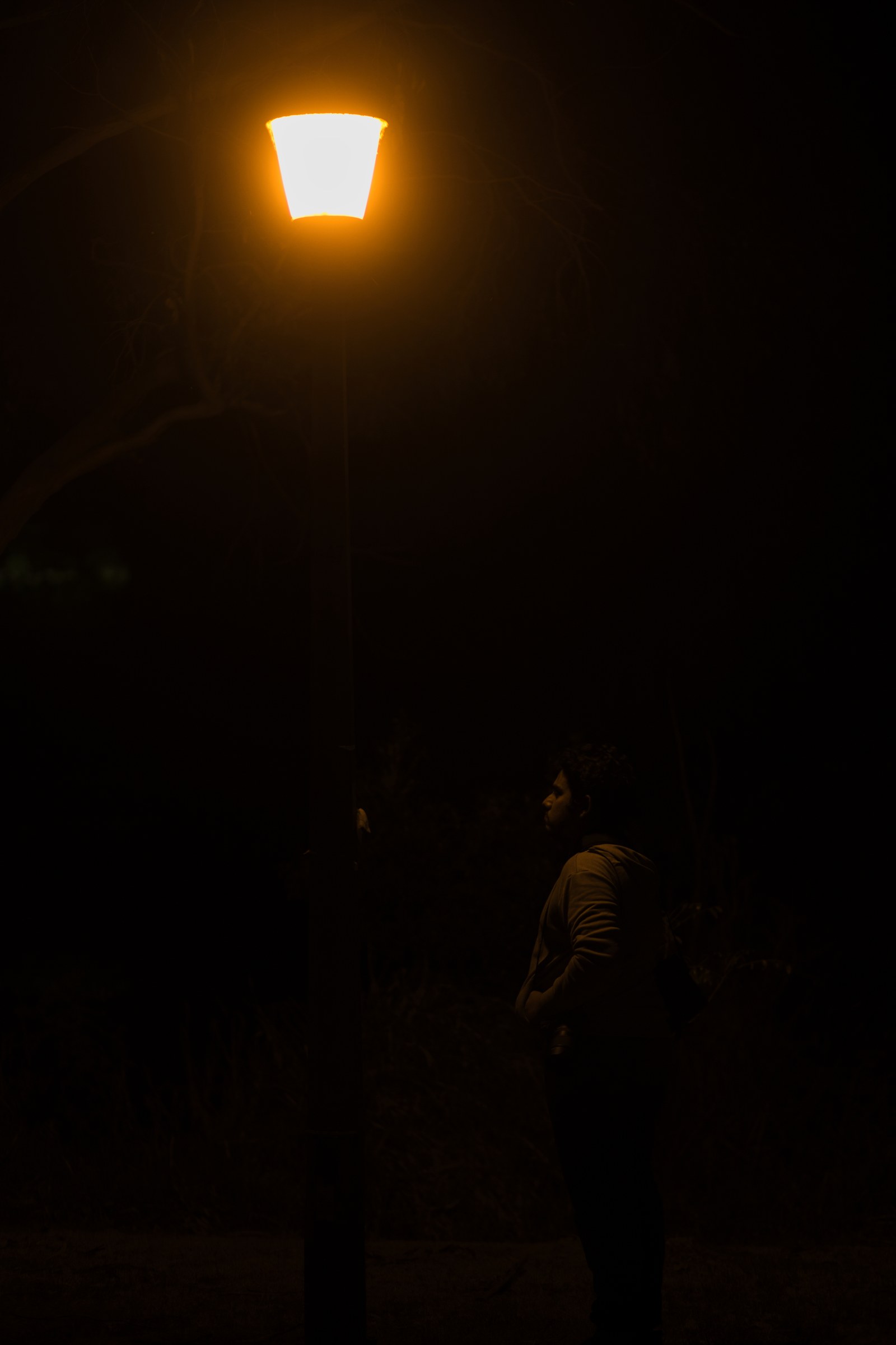 Devindu Gamage stands in deep shadow beneath a glowing streetlamp, shown in a vertical night portrait.