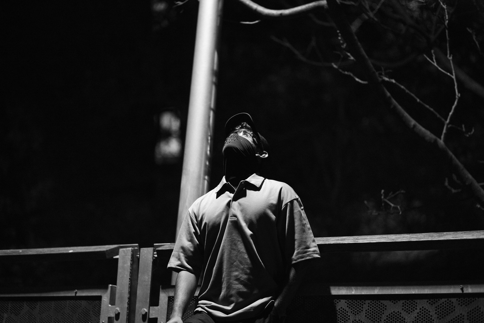 A monochrome portrait of a person leaning back beneath a streetlamp beside a railing.