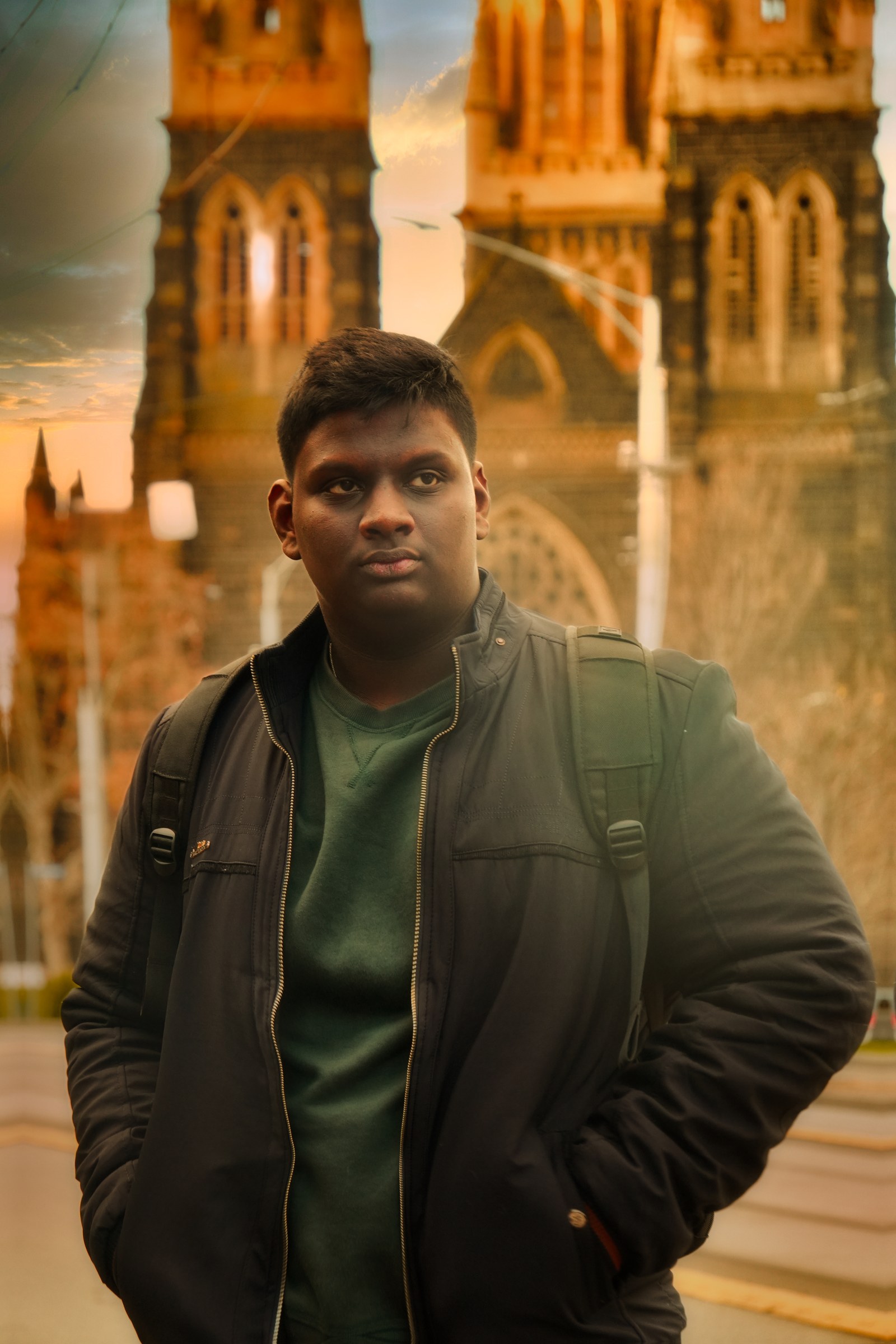 A person in a dark jacket stands in front of a stone building with tall towers.
