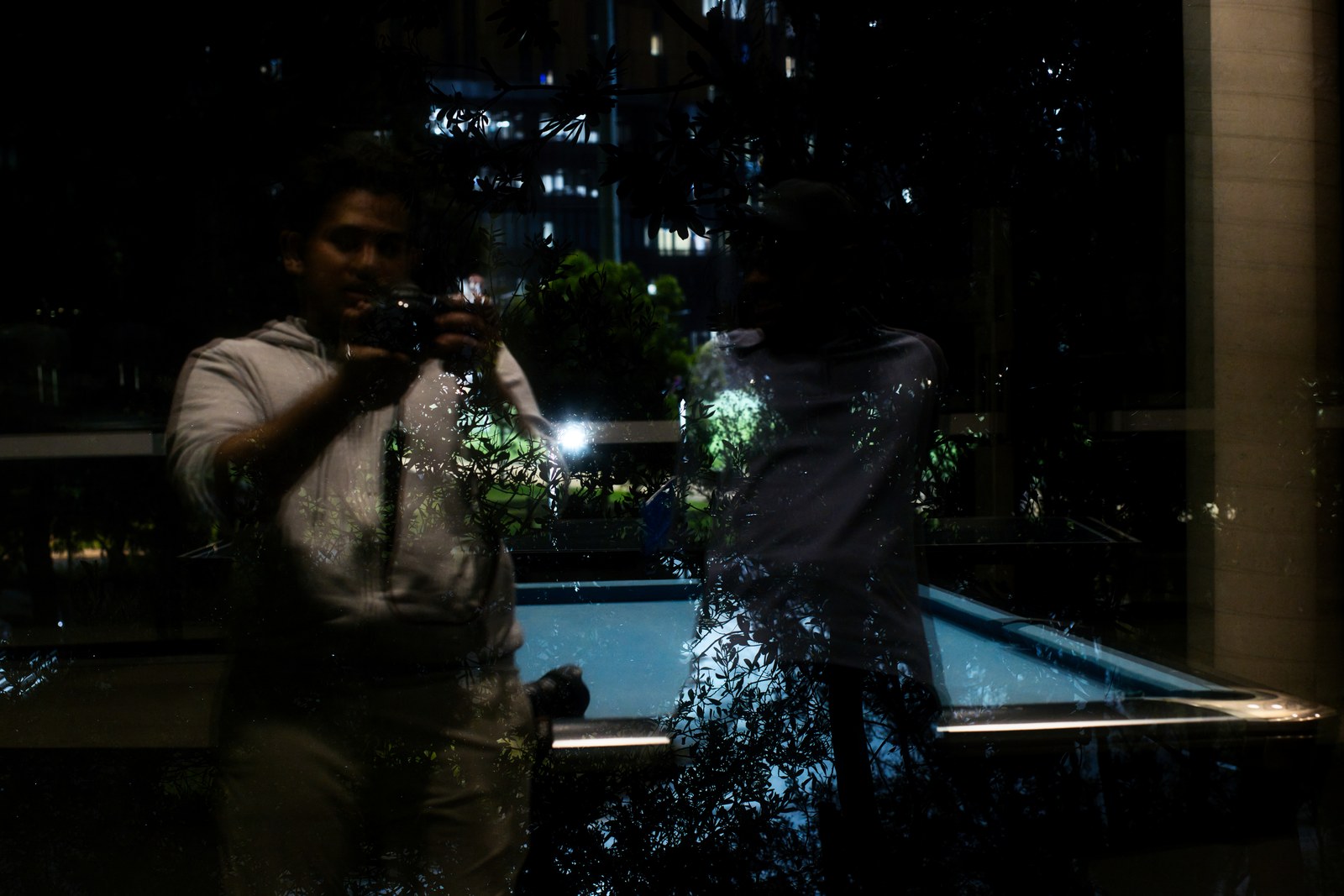 A person holding a camera is reflected in dark glass beside a lit pool at night.