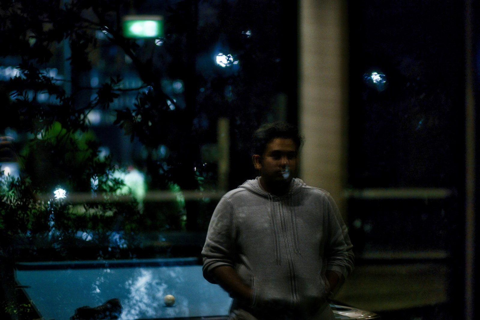 A person stands in dim light beside reflective glass and a pool at night.