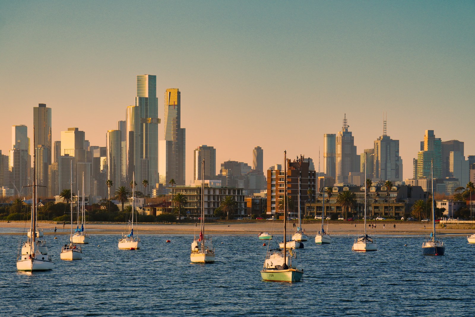 Moored sailing boats fill the water below a city skyline in warm evening light.