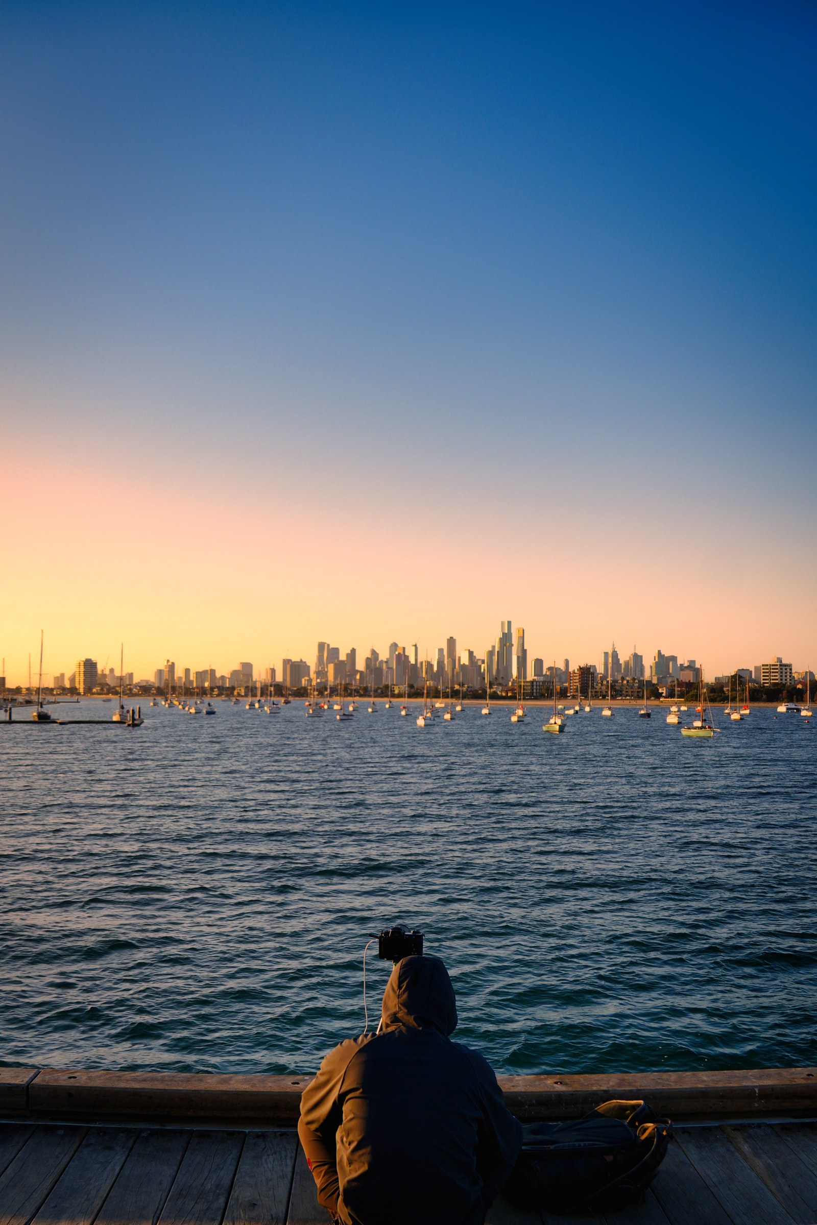 A seated person faces water, moored boats and a distant skyline beneath a blue and pink sky.