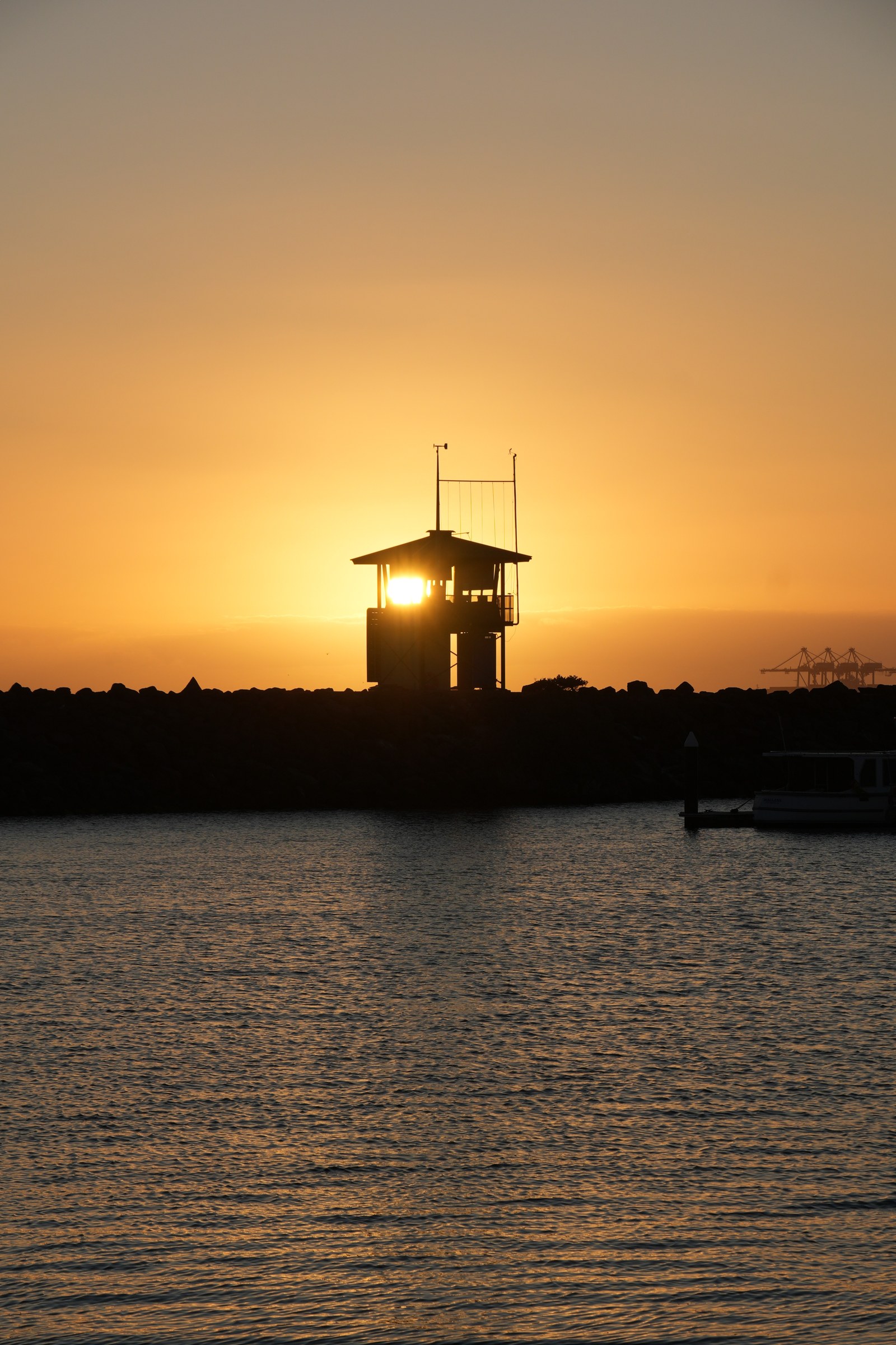 Golden sunlight shines through a small tower on a dark breakwater above calm water.