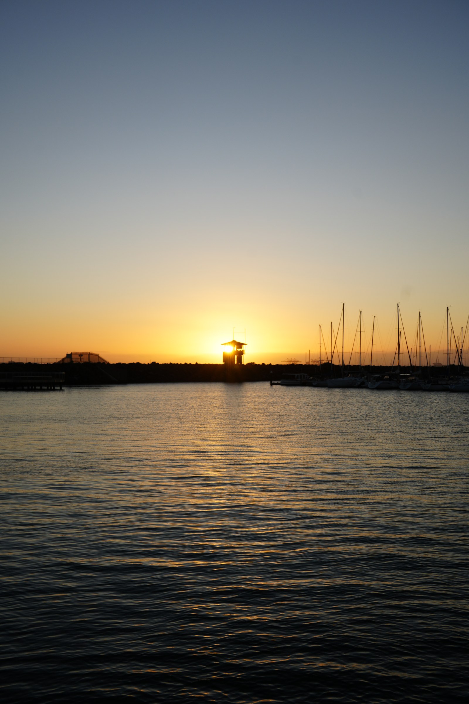 A wide evening sky rises above water, with the sun low behind a waterfront tower and yacht masts.