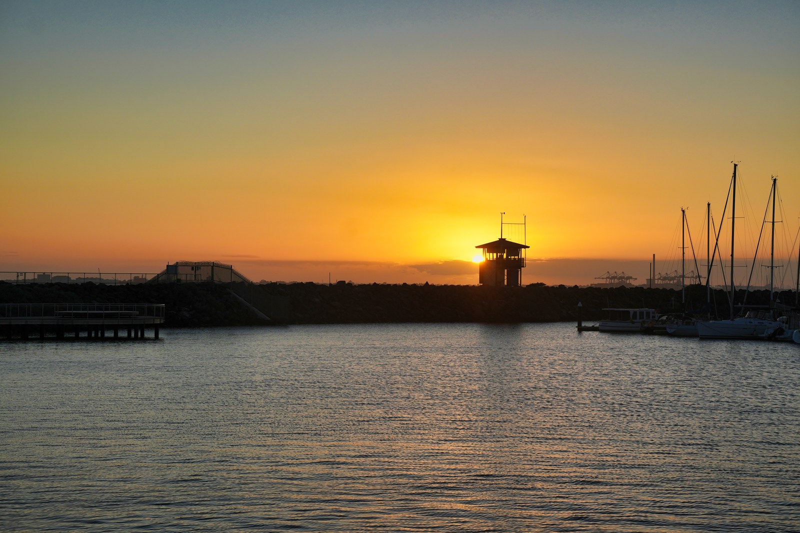 The low sun glows behind a small waterfront tower beside the silhouettes of yacht masts.